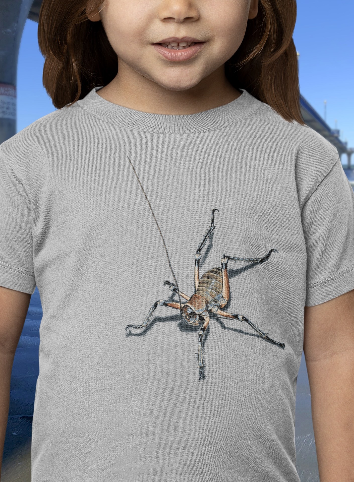 Close-up of a little girl smiling, wearing a grey marle t-shirt with an all-over weta print, with New Brighton Pier in the background.