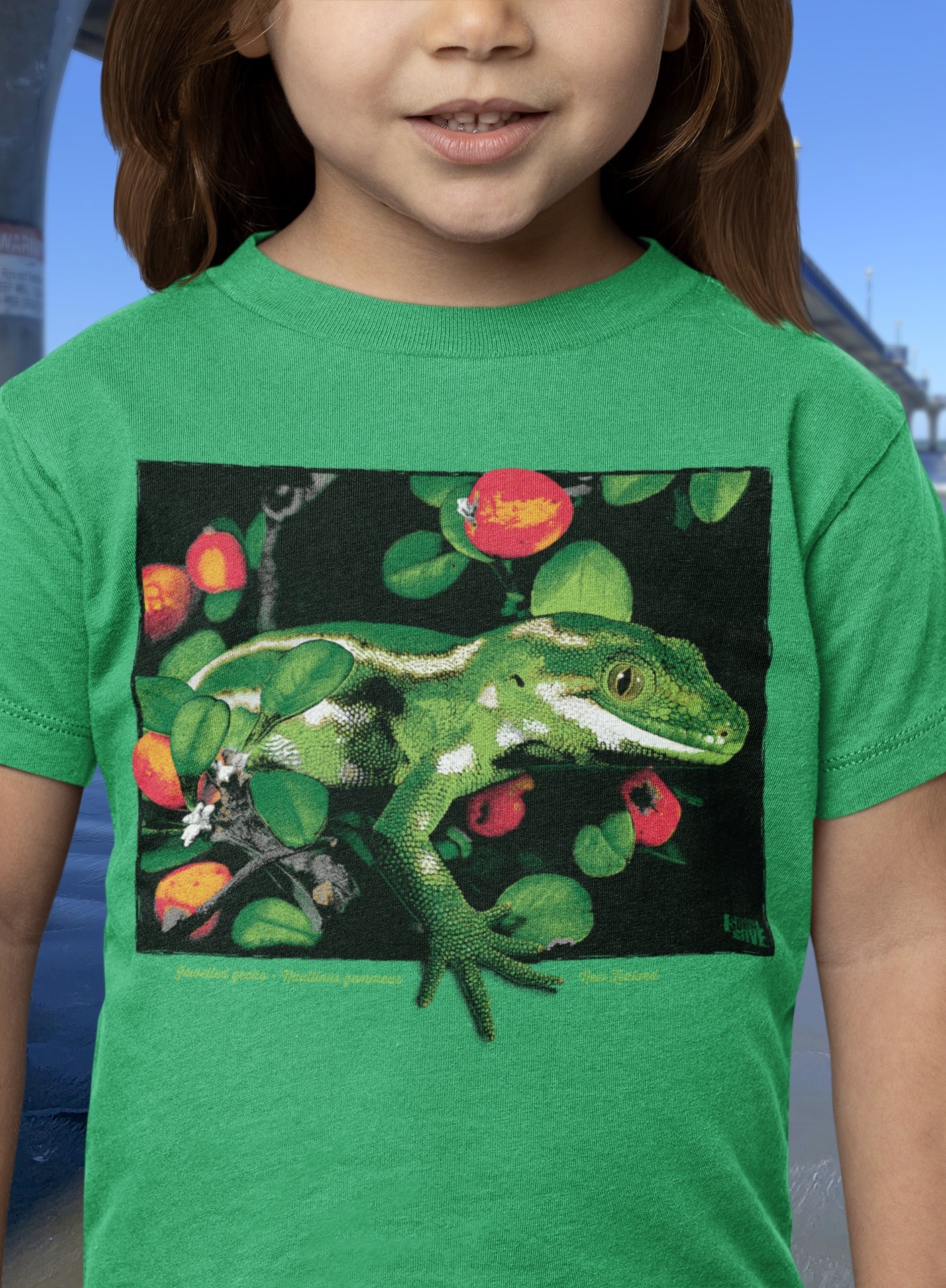 A young girl in a Kelly green t-shirt with a jewelled gecko print stands smiling by New Brighton Pier, showcasing the shirt's vibrant design.