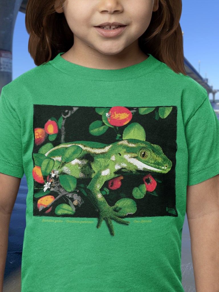 A young girl in a Kelly green t-shirt with a jewelled gecko print stands smiling by New Brighton Pier, showcasing the shirt's vibrant design.