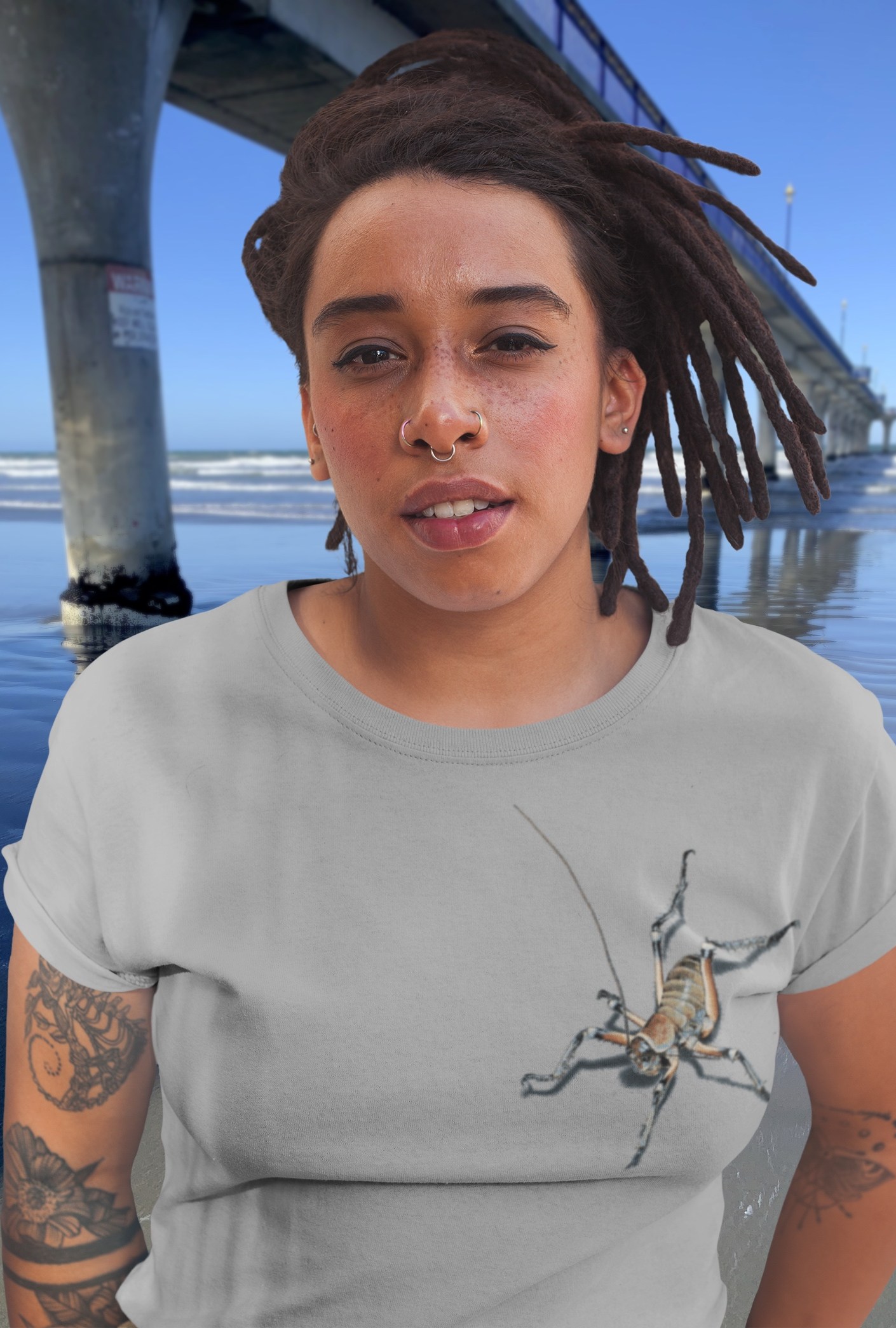 A woman with braids in a grey marle t-shirt featuring an all-over weta print stands beside New Brighton Pier, with the Pacific ocean in the background.