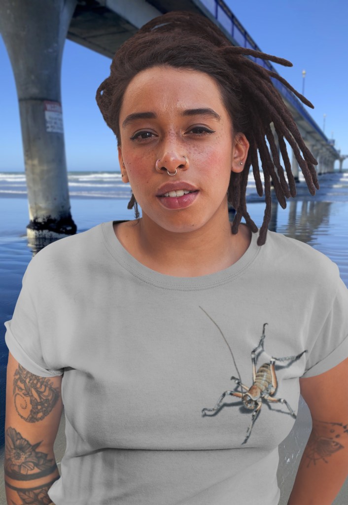 A woman with braids in a grey marle t-shirt featuring an all-over weta print stands beside New Brighton Pier, with the Pacific ocean in the background.