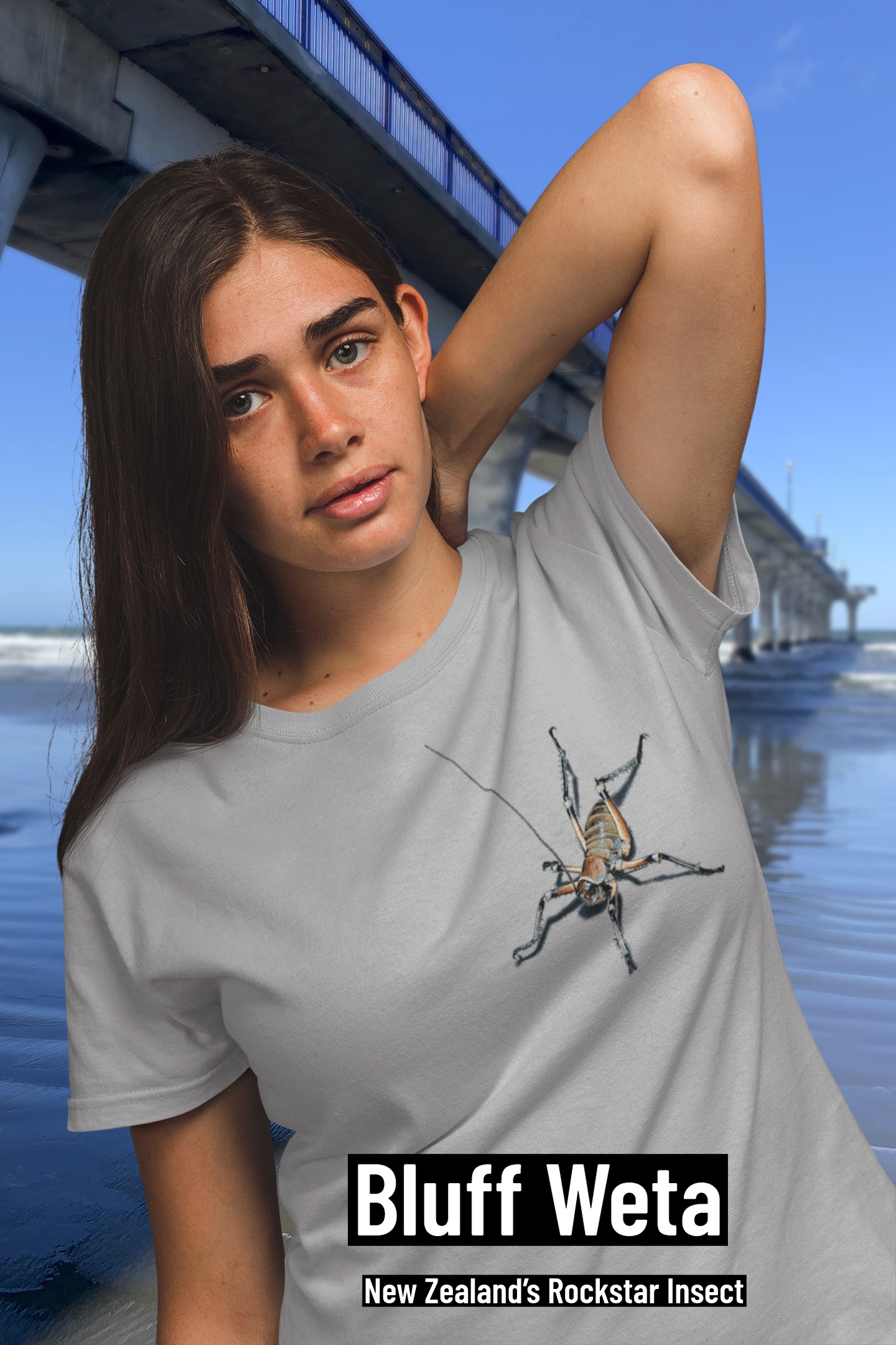 A young woman with long black hair wears a grey marle t-shirt with an all-over weta print, posing by New Brighton Pier.