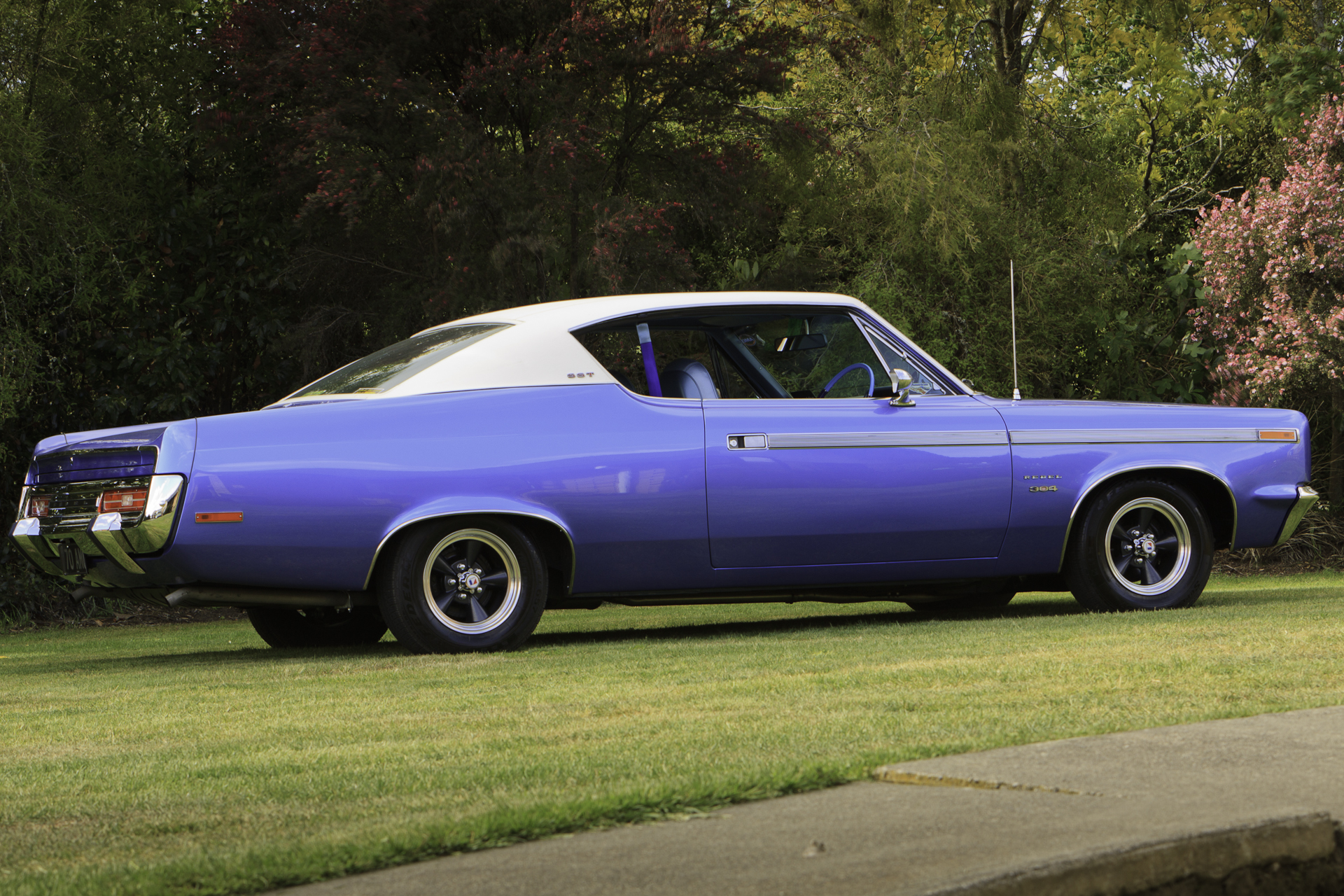 Low-angle side view of a royal blue 1970 Rambler Rebel SST showcasing the rear bumper and tail lights, highlighting the car's restored elegance.