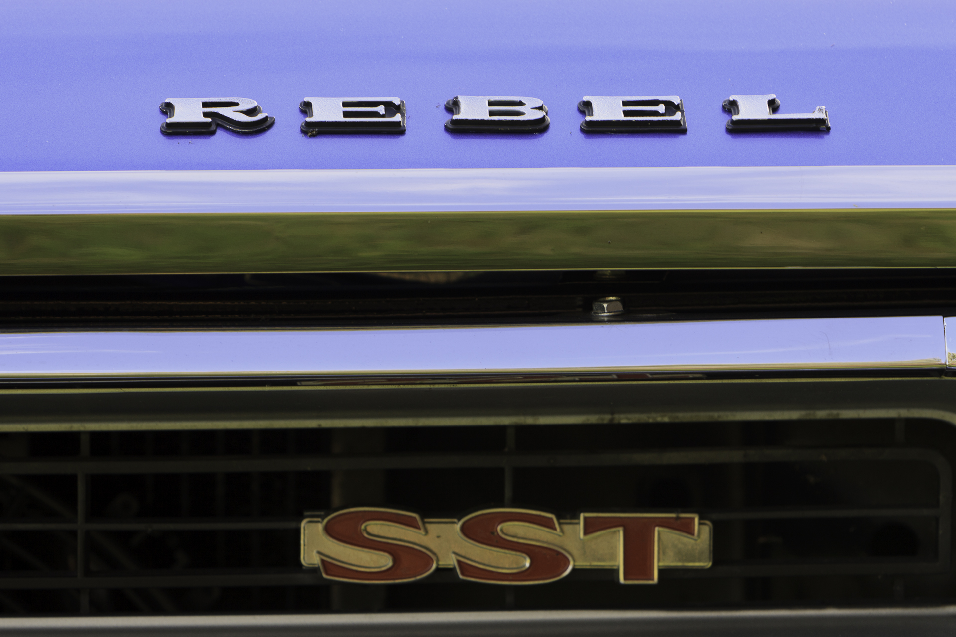 Close-up of the 'Rebel - SST' badge on the grille of a royal blue 1970 Rambler Rebel SST, emphasizing the car's classic muscle identity