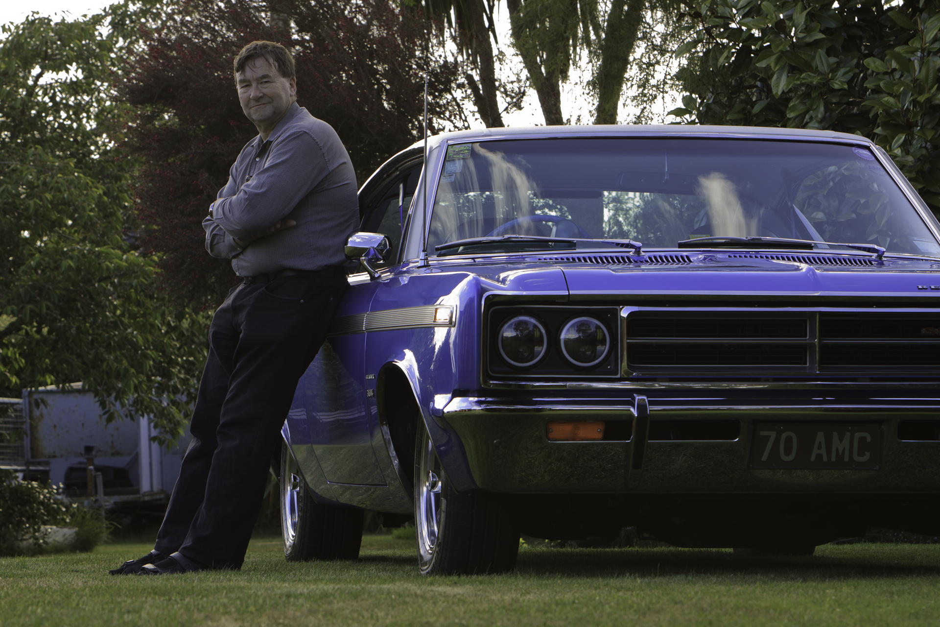 Lindsay McKenzie, owner and restoration project manager, with his restored 1970 Rambler Rebel SST in royal blue, leaning against the car's driver's door.