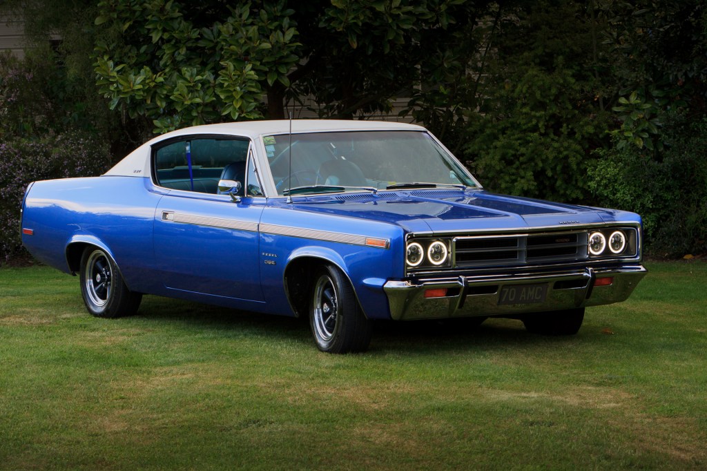 Royal blue 1970 Rambler Rebel SST Hardtop Coupe against a New Zealand garden landscape, highlighting its elegant design and chrome details.