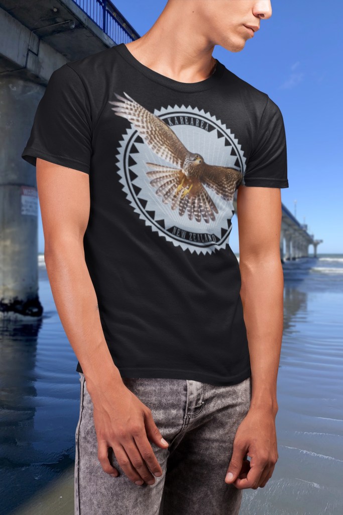 A man beside New Brighton Pier wears a black t-shirt with an asymmetric print of a karearea in flight, wings extending onto the sleeve and wrapping around to the back.