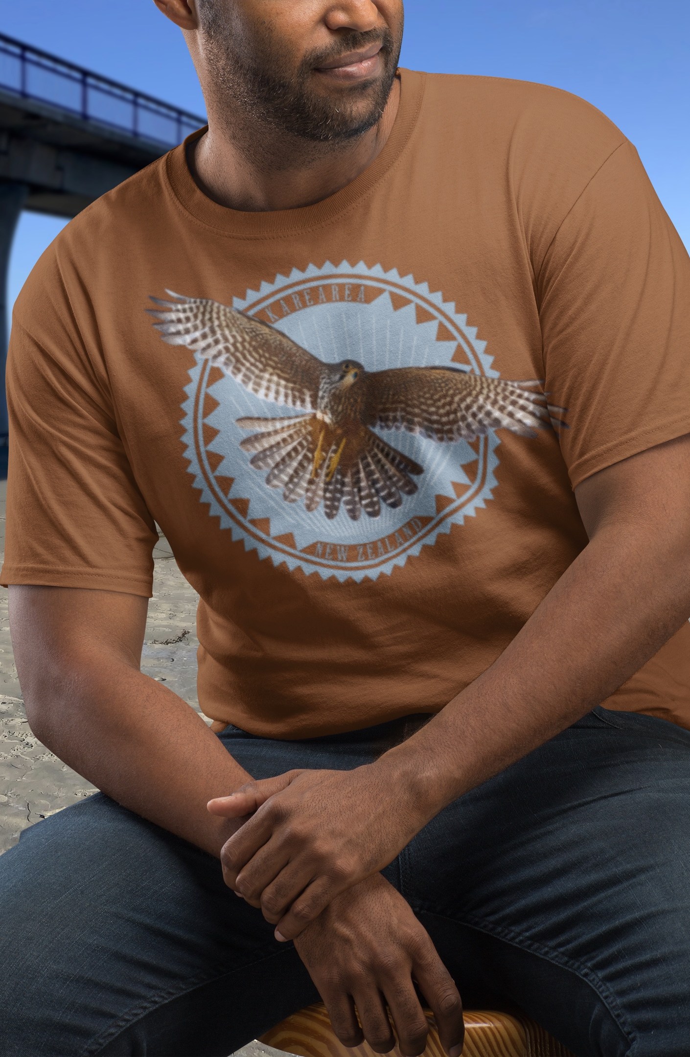 A man wearing a clay-coloured t-shirt with a karearea (New Zealand falcon) in flight print, seated next to New Brighton Pier.