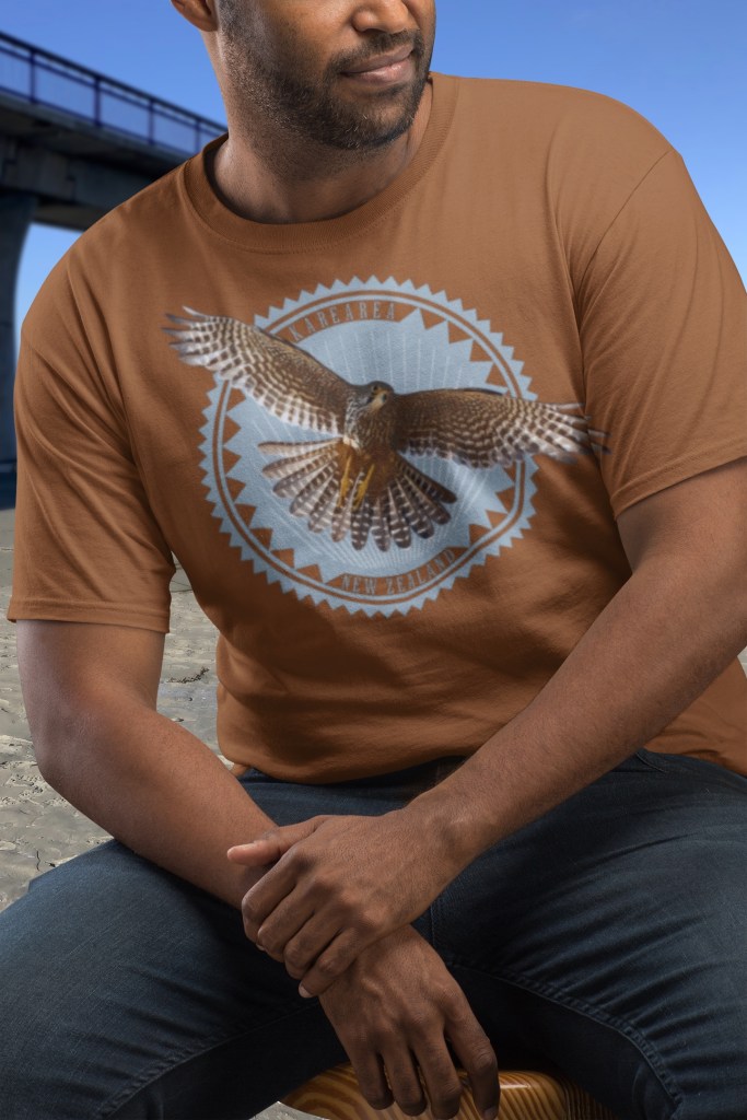 A man wearing a clay-coloured t-shirt with a karearea (New Zealand falcon) in flight print, seated next to New Brighton Pier.