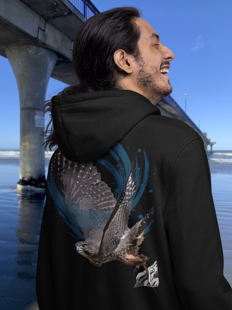 A laughing man with long hair wearing a black hooded sweatshirt with a large artistic print of a New Zealand falcon (karearea) in flight on the back, photographed on the beach beside New Brighton Pier in Christchurch, New Zealand. The design features the detailed falcon with spread wings against swirling blue abstract brushstrokes on a black hoodie background.