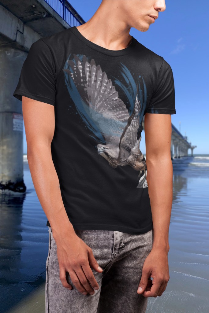 A man wearing a black T-shirt with a bold artistic print of a New Zealand falcon (karearea) taking flight, photographed on the beach beside New Brighton Pier in Christchurch, New Zealand. The design shows the detailed falcon with outstretched wings against swirling blue abstract brushstrokes on a black shirt background.