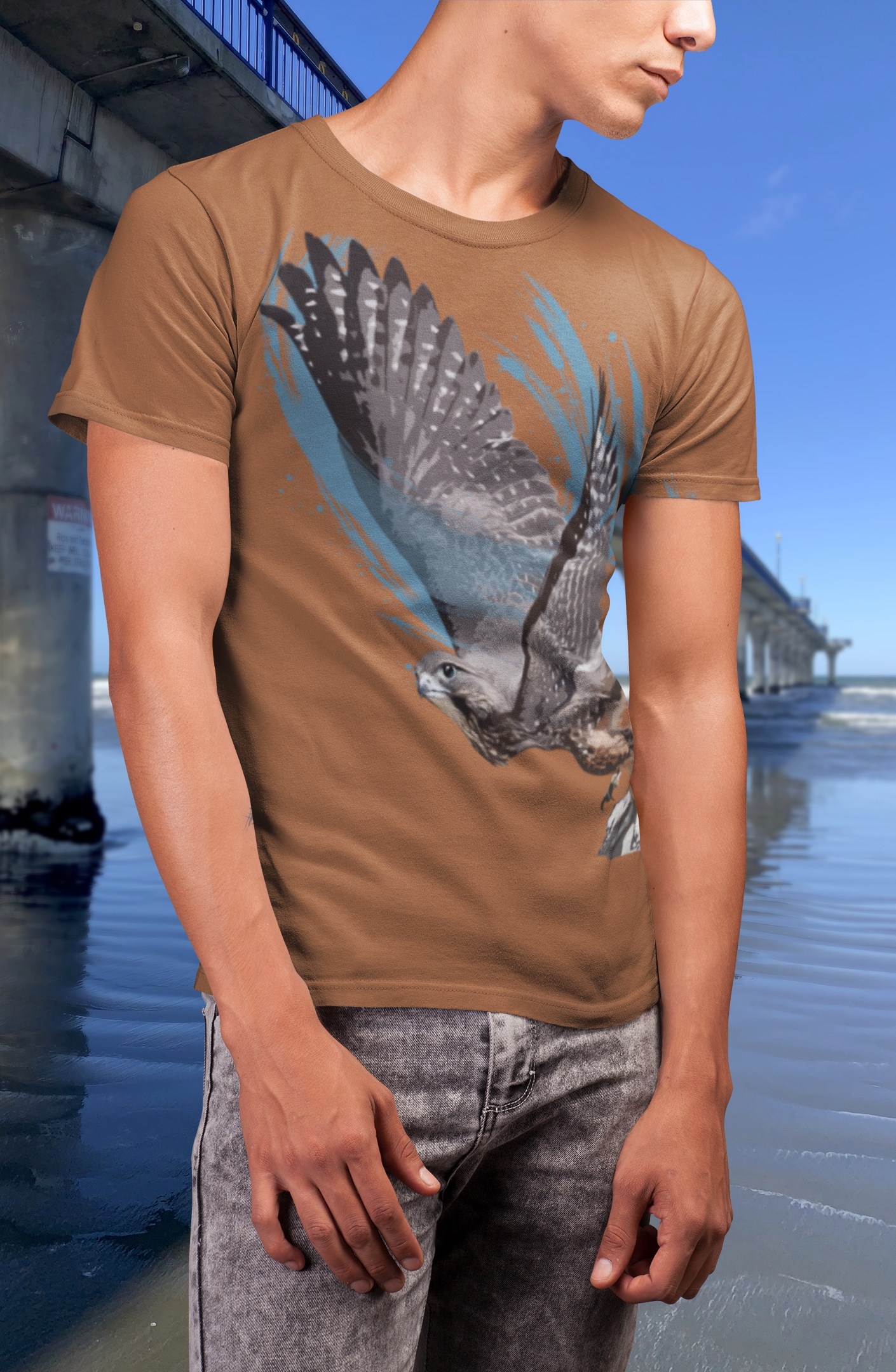 A man wearing a rust-brown T-shirt with a bold artistic print of a New Zealand falcon (kārearea) soaring in flight, photographed on the beach beside New Brighton Pier in Christchurch, New Zealand. The design features detailed falcon wings against blue abstract brushstrokes on a warm earthy shirt background.