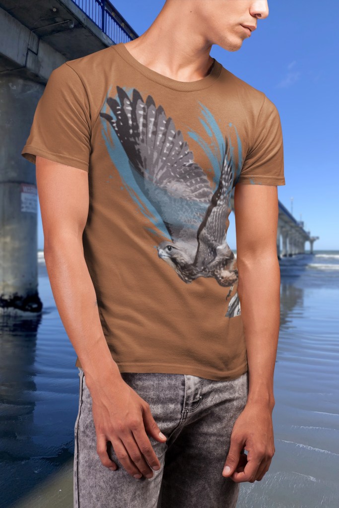 A man wearing a rust-brown T-shirt with a bold artistic print of a New Zealand falcon (kārearea) soaring in flight, photographed on the beach beside New Brighton Pier in Christchurch, New Zealand. The design features detailed falcon wings against blue abstract brushstrokes on a warm earthy shirt background.
