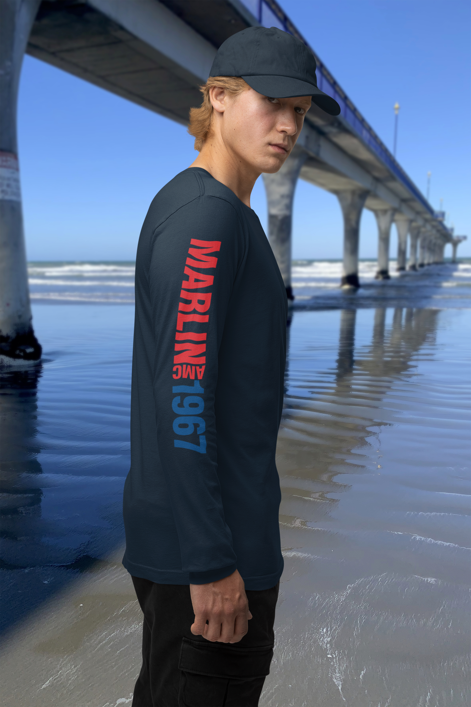 Man wearing navy blue long-sleeve t-shirt with 1967 AMC Marlin typographic arm print, standing by New Brighton Pier.
