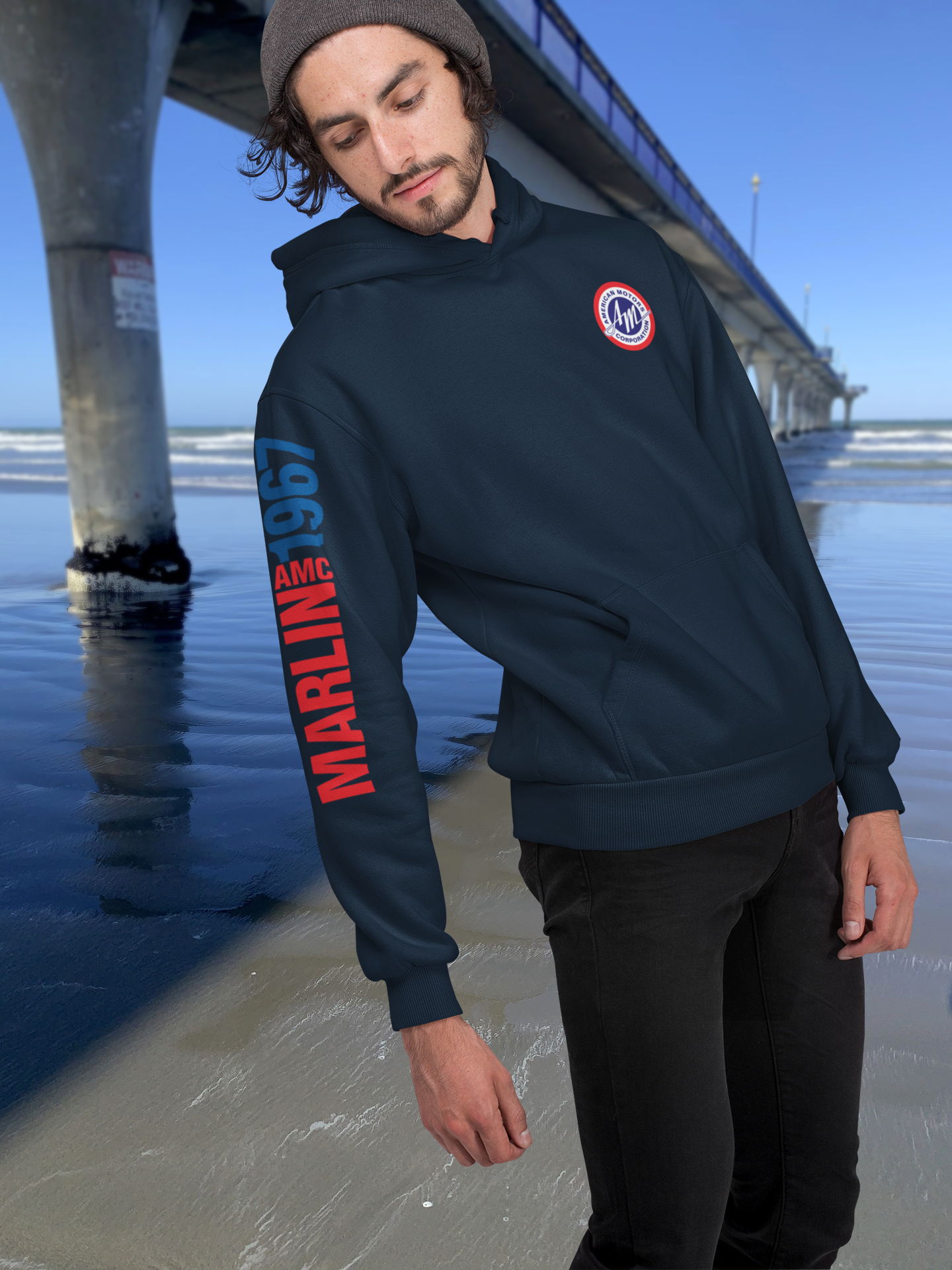 Man in navy blue 1967 AMC Marlin hoodie with round American Motors logo, standing beside New Brighton's concrete pier under a clear blue sky.