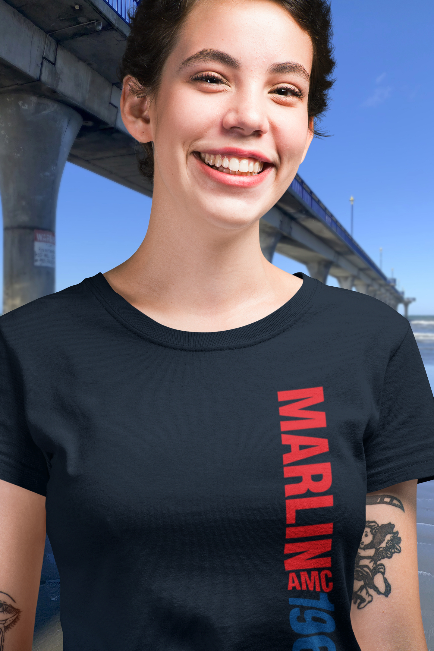 Smiling woman in navy blue t-shirt with 1967 AMC Marlin text, beside New Brighton's concrete pier under a clear blue sky.