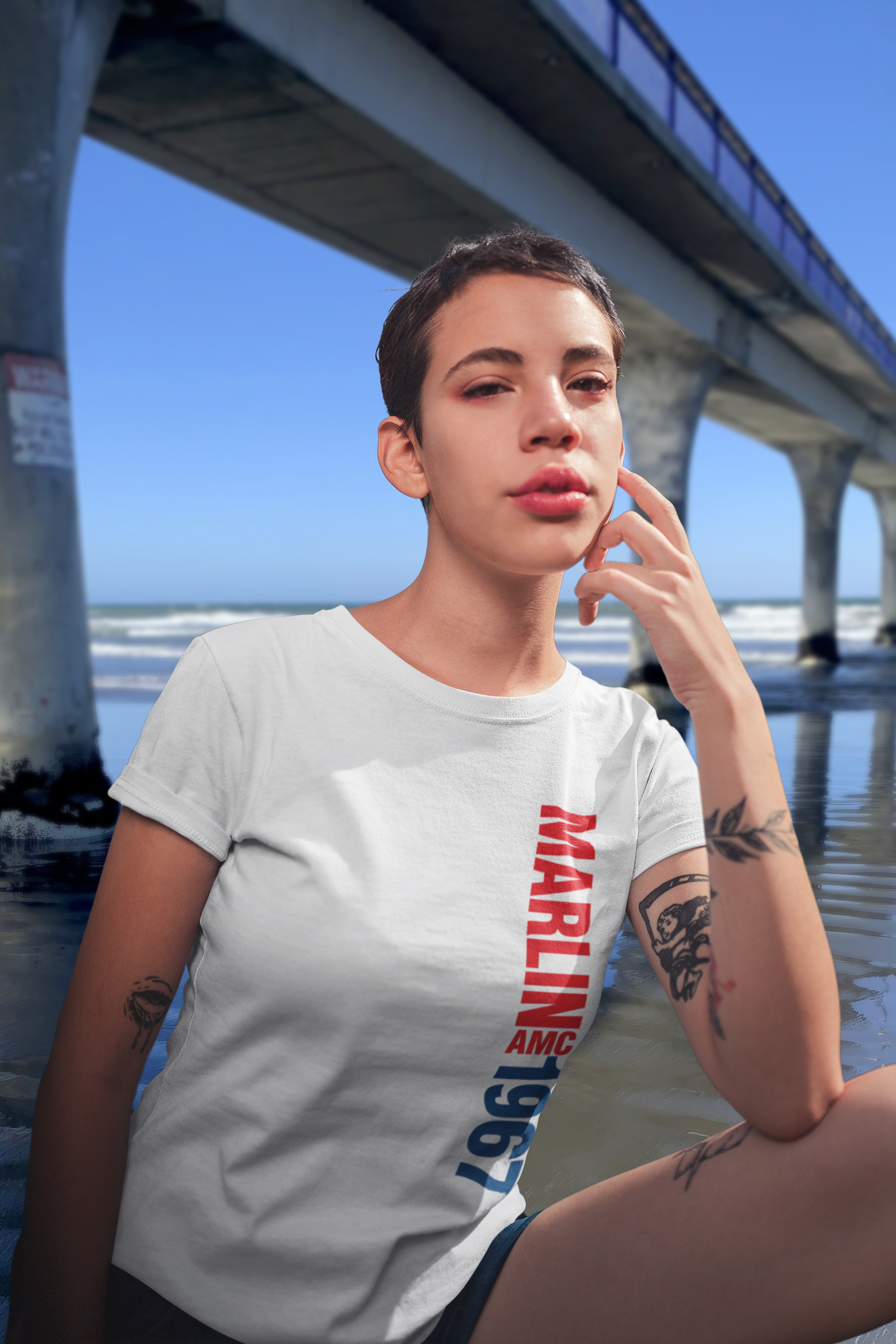 Woman wearing white t-shirt with 1967 AMC Marlin typography, standing beside the concrete New Brighton Pier with blue sky background.