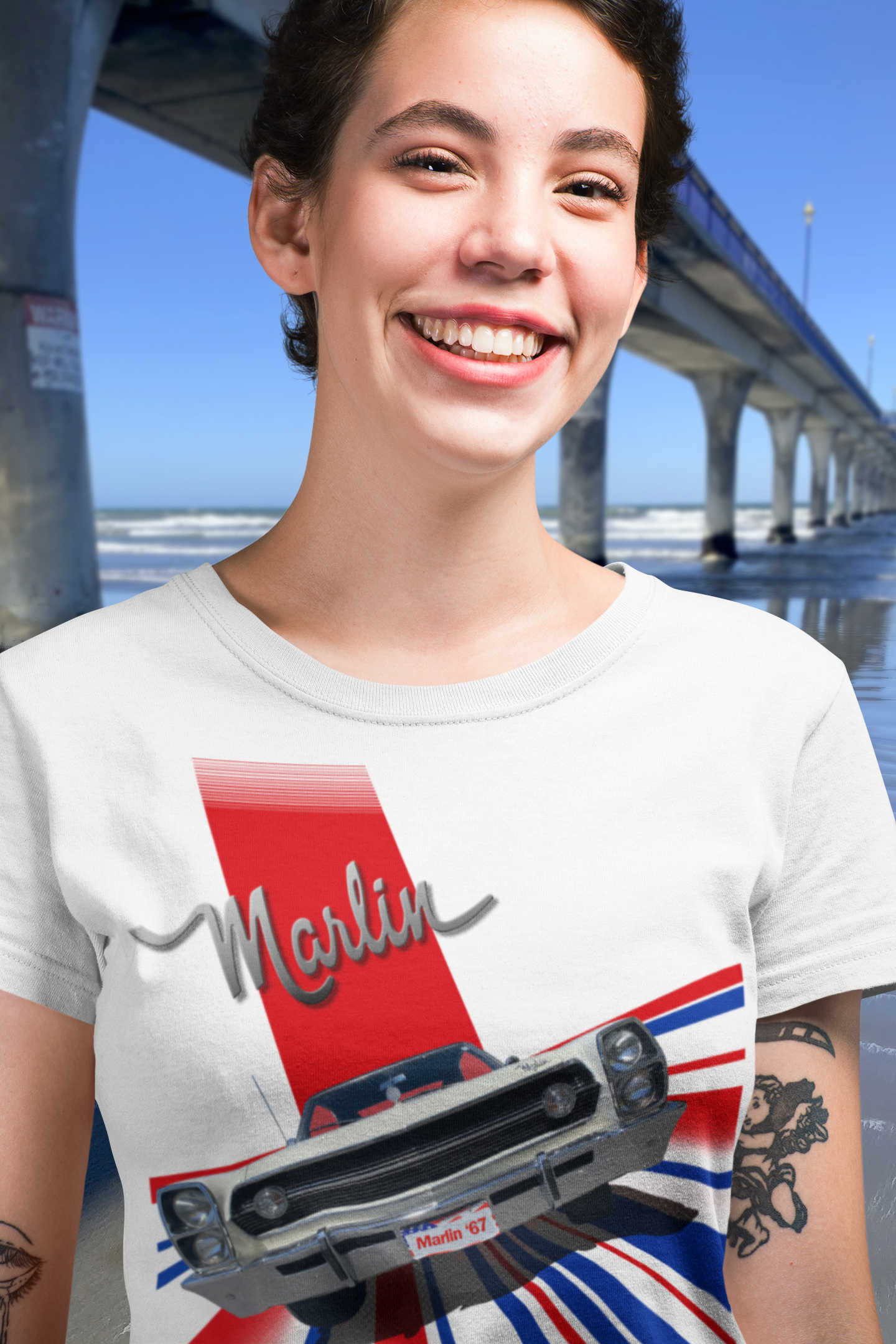 Woman in white t-shirt with 1967 AMC Marlin front view design, beside New Brighton Pier under a clear blue sky.