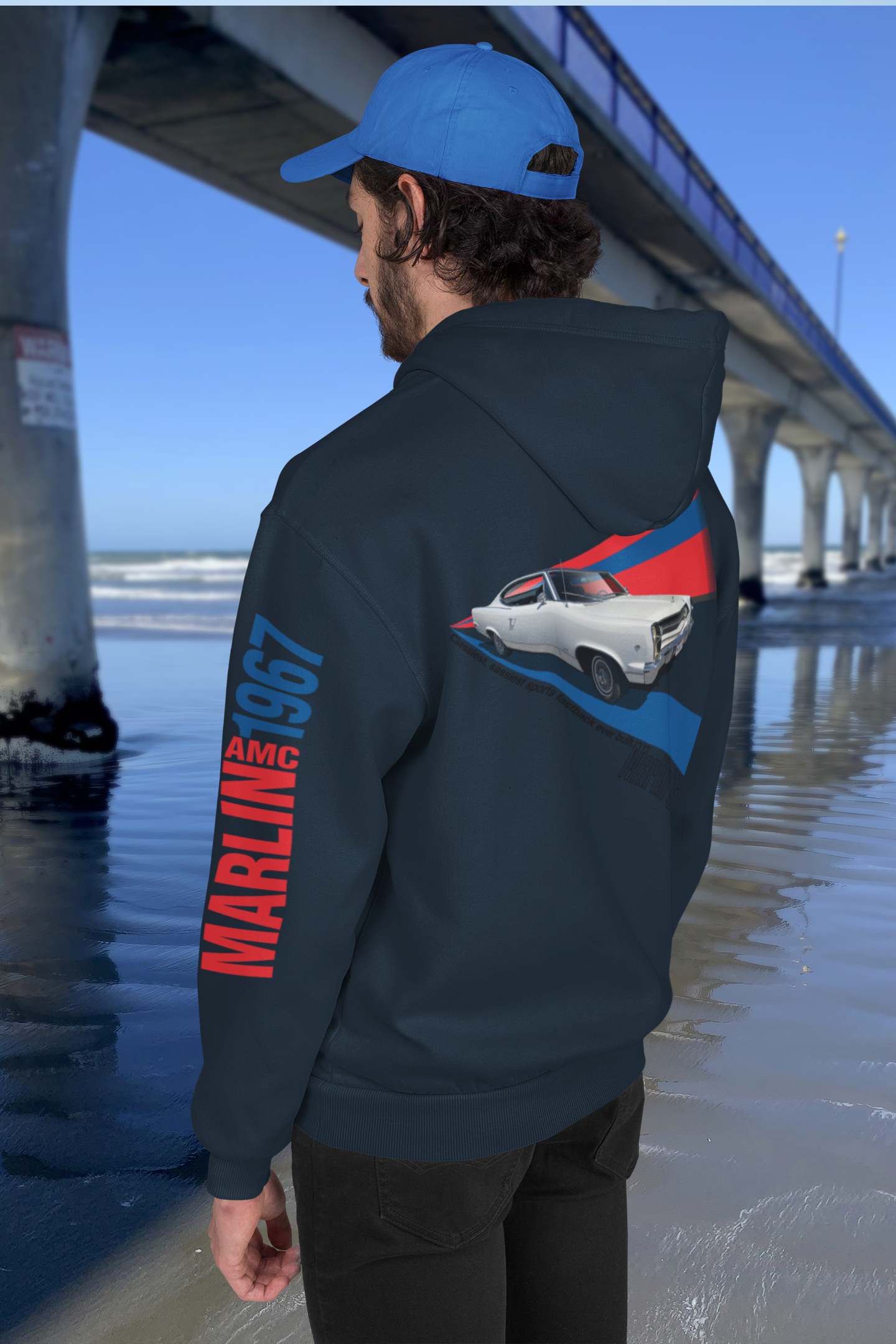 Man wearing navy blue hoodie with 1967 AMC Marlin graphic beside New Brighton's concrete pier under a clear blue sky.