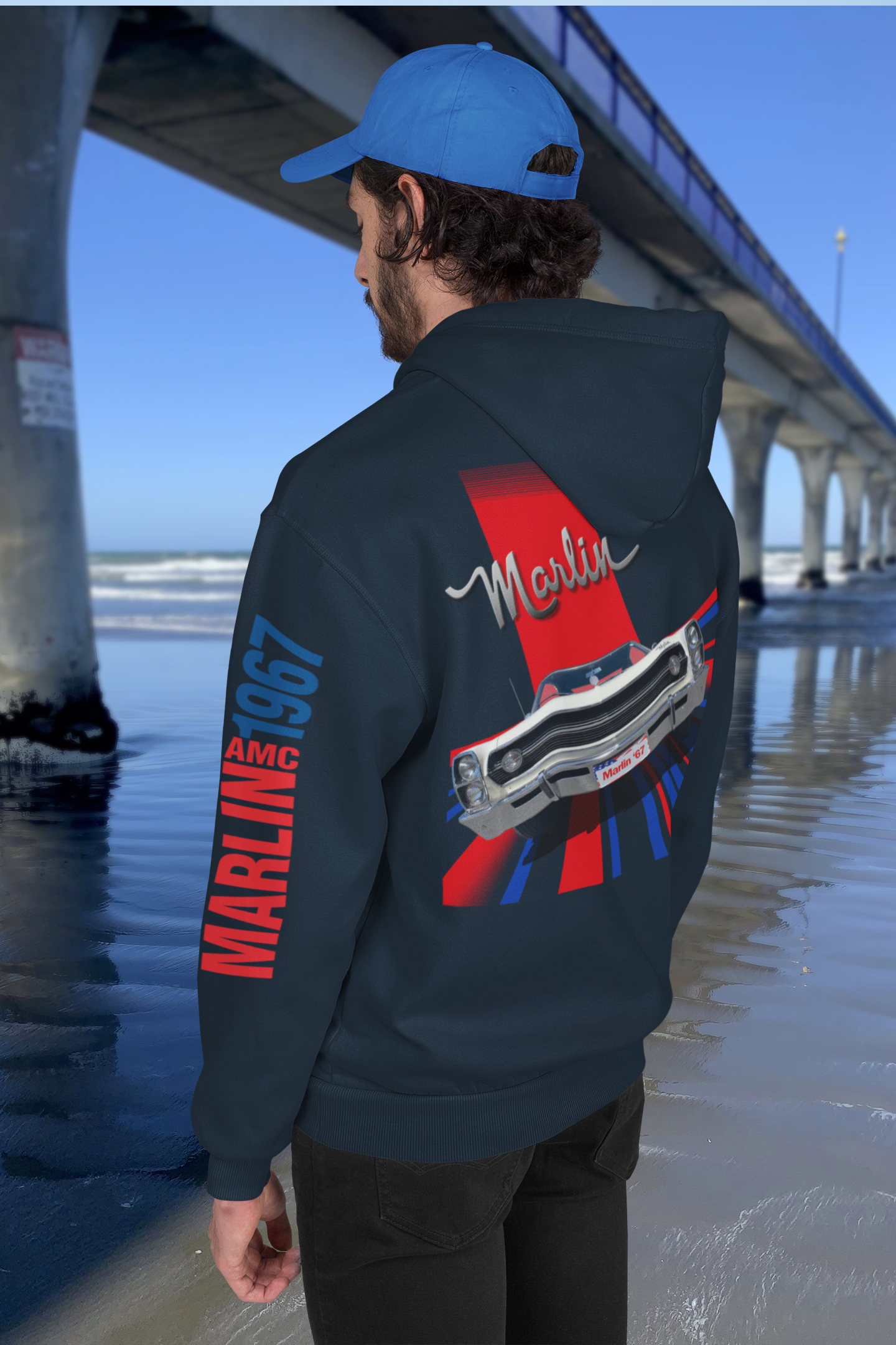Back three-quarter view of woman in navy blue hoodie with 1967 AMC Marlin front view graphic, standing by New Brighton's concrete pier under clear blue sky.
