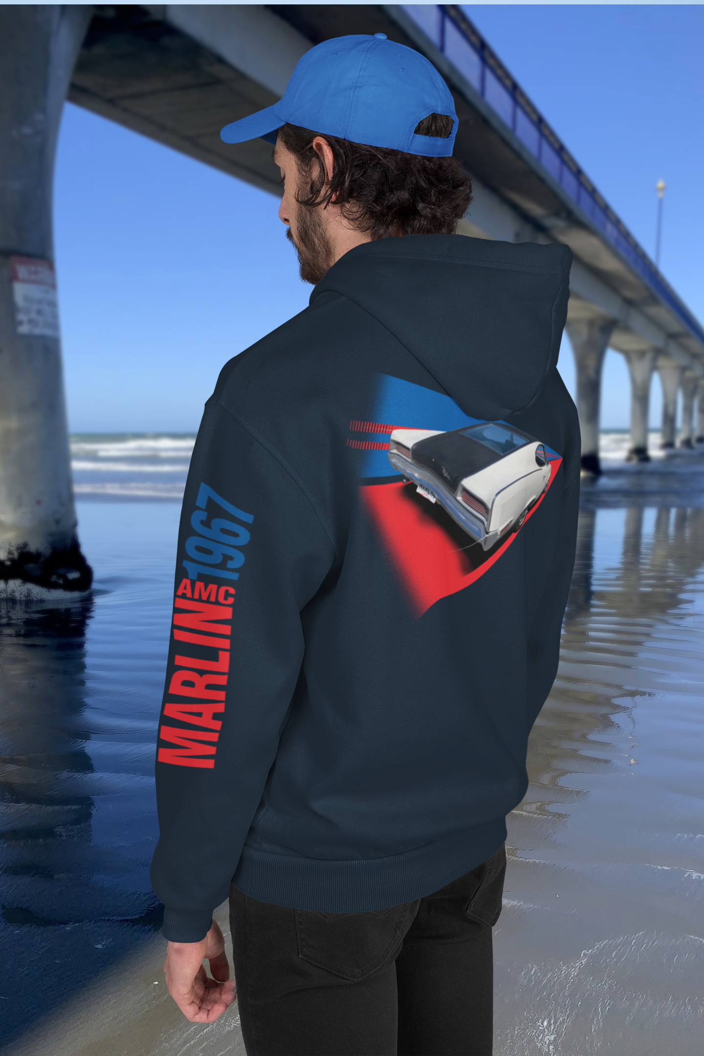 Man in navy blue 1967 AMC Marlin sport fastback hoodie beside New Brighton's concrete pier under a clear blue sky.