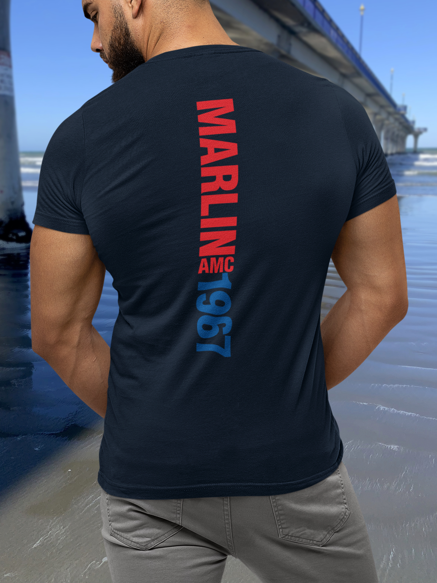 Back view of a man wearing navy blue t-shirt with 1967 AMC Marlin typography, standing by New Brighton's concrete pier under a clear blue sky.