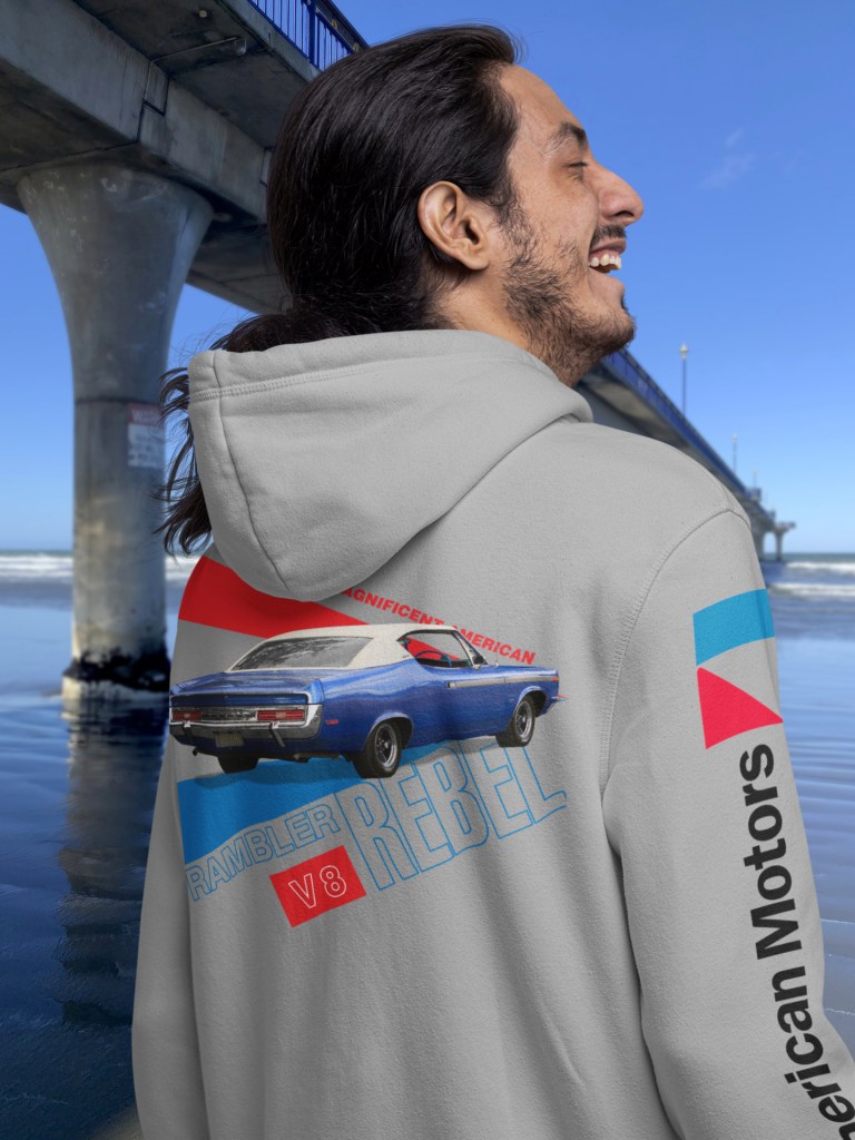 Man wearing grey marle hoodie with 1970 Rambler Rebel SST print, standing by New Brighton Pier, New Zealand.