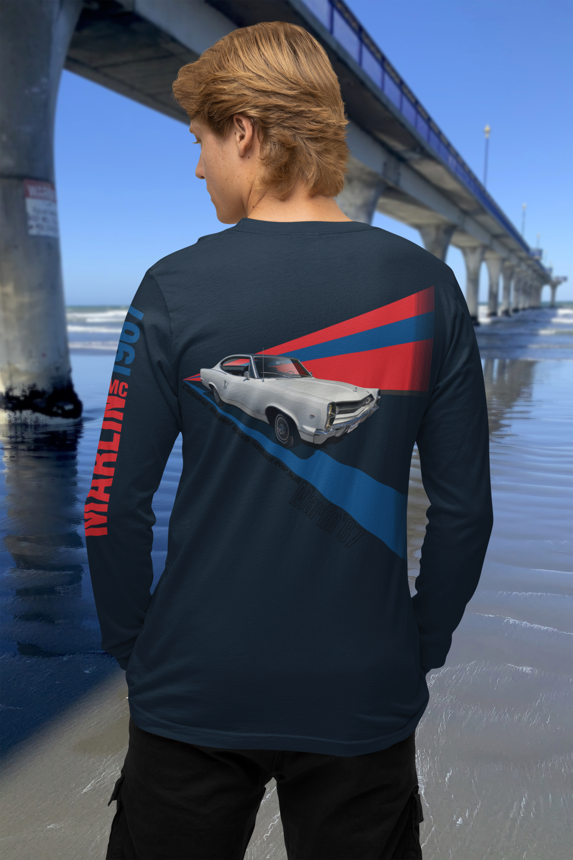 Back view of a man wearing a navy blue t-shirt with a 1967 AMC Marlin graphic, beside New Brighton Pier.