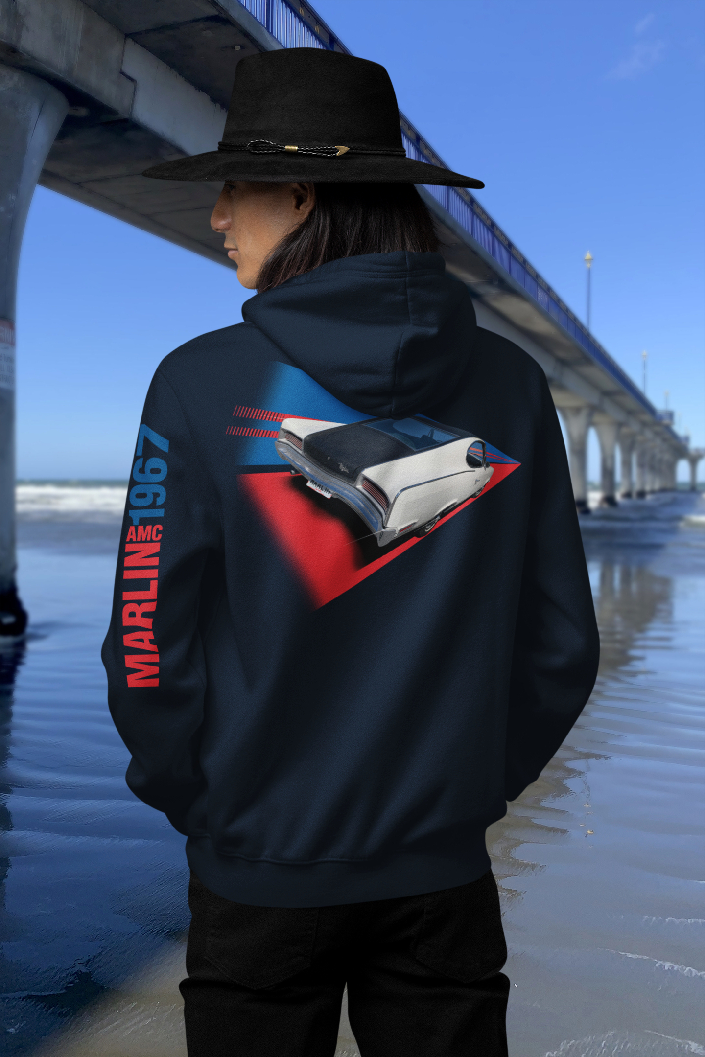 Person wearing a navy blue hoodie with 1967 AMC Marlin Sport Fastback print, standing in front of New Brighton Pier with a clear blue sky.