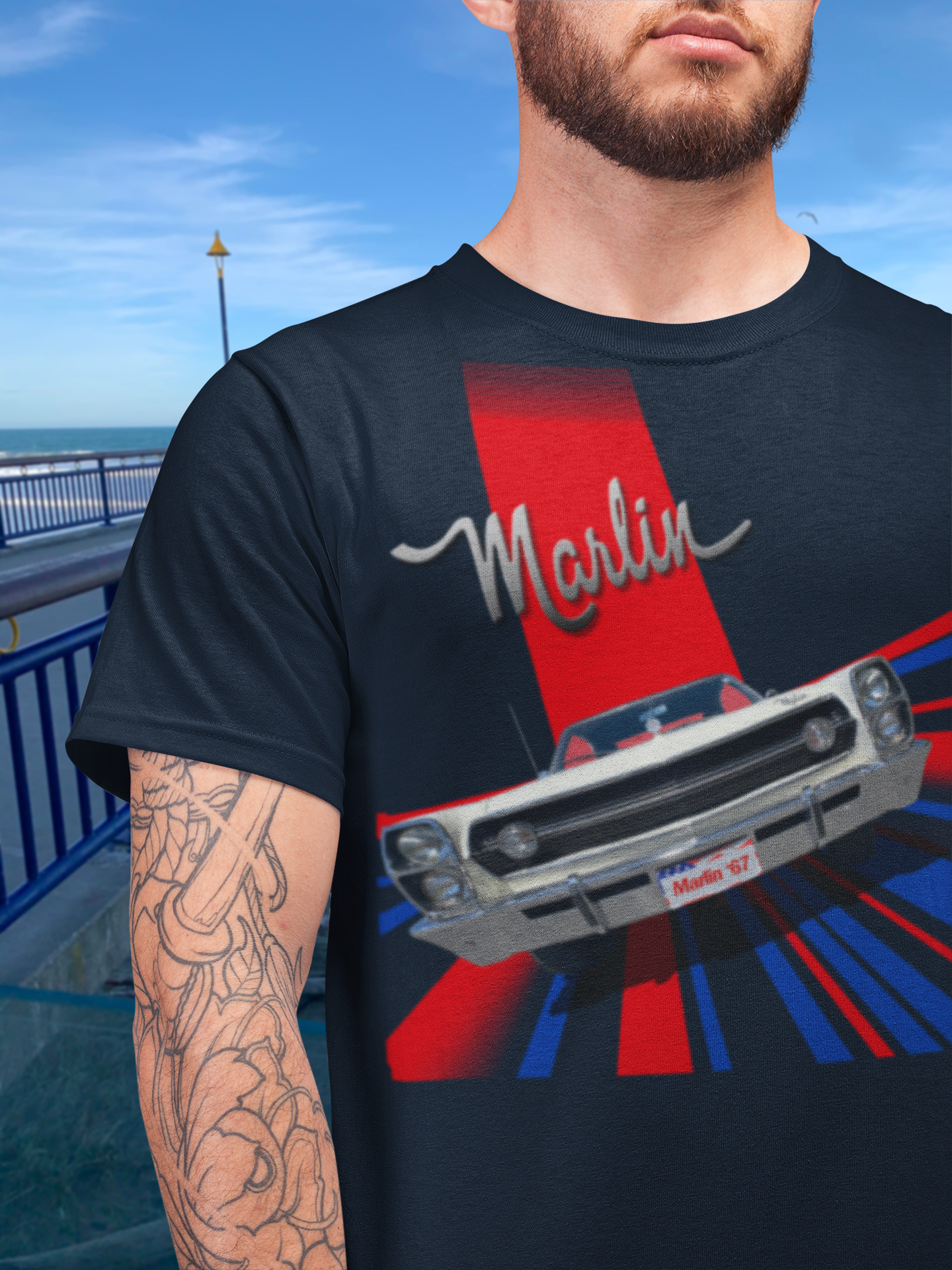 Bearded man wearing a navy blue t-shirt featuring a 1967 AMC Marlin in a 1960s Grand Prix poster style, beside New Brighton Pier.