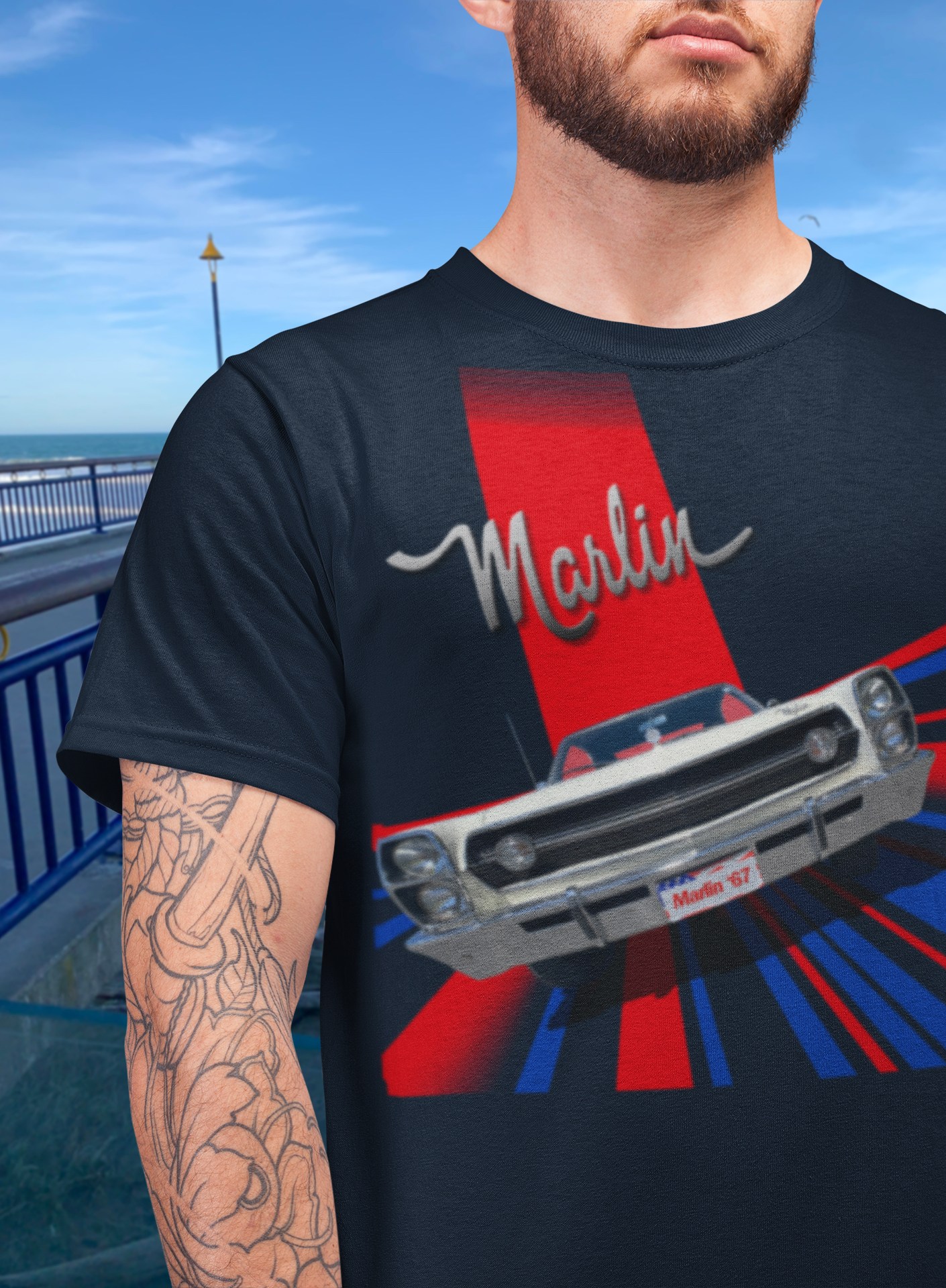 Bearded man wearing a navy blue t-shirt featuring a 1967 AMC Marlin in a 1960s Grand Prix poster style, beside New Brighton Pier.
