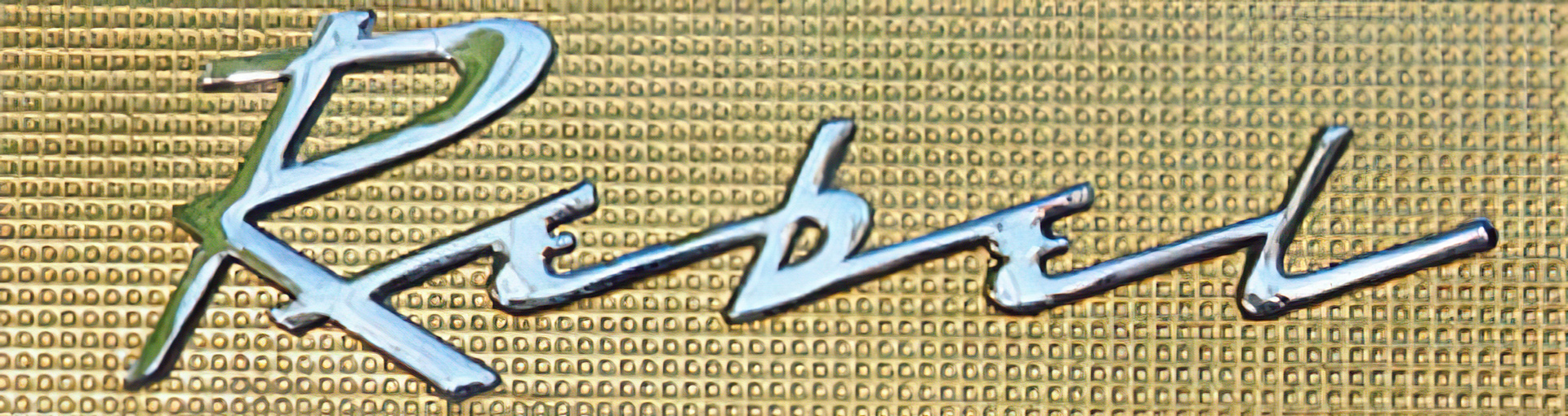 Close-up of the chrome ‘Rebel’ name badge on a 1957 Rambler Rebel sedan, set against textured gold-anodised aluminum.