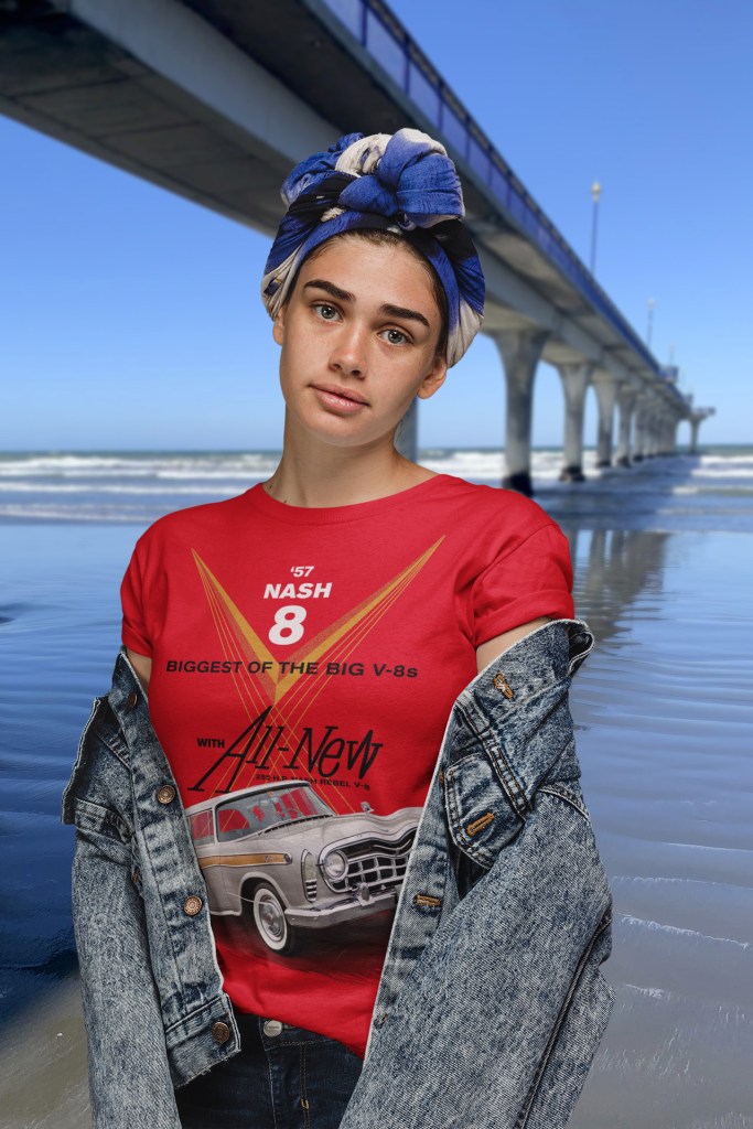 Front view of a young woman in a red t-shirt with a 1957 Rambler Rebel design, including a test track illustration of the car in motion and a small badge print on the back, beside New Brighton Pier.