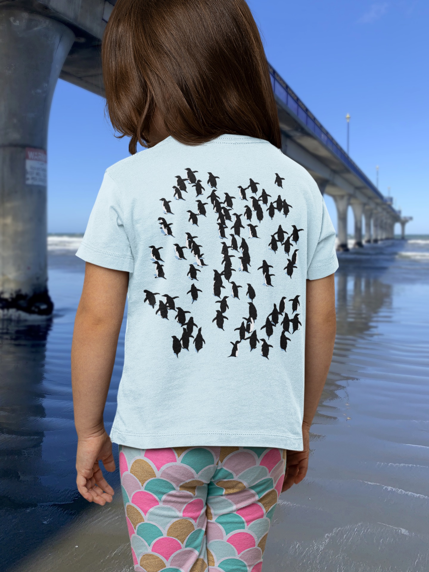 Back view of a young girl in a light blue Adelie Penguin t-shirt beside New Brighton Pier, showing penguins fleeing a leopard seal in black, white, and blue.