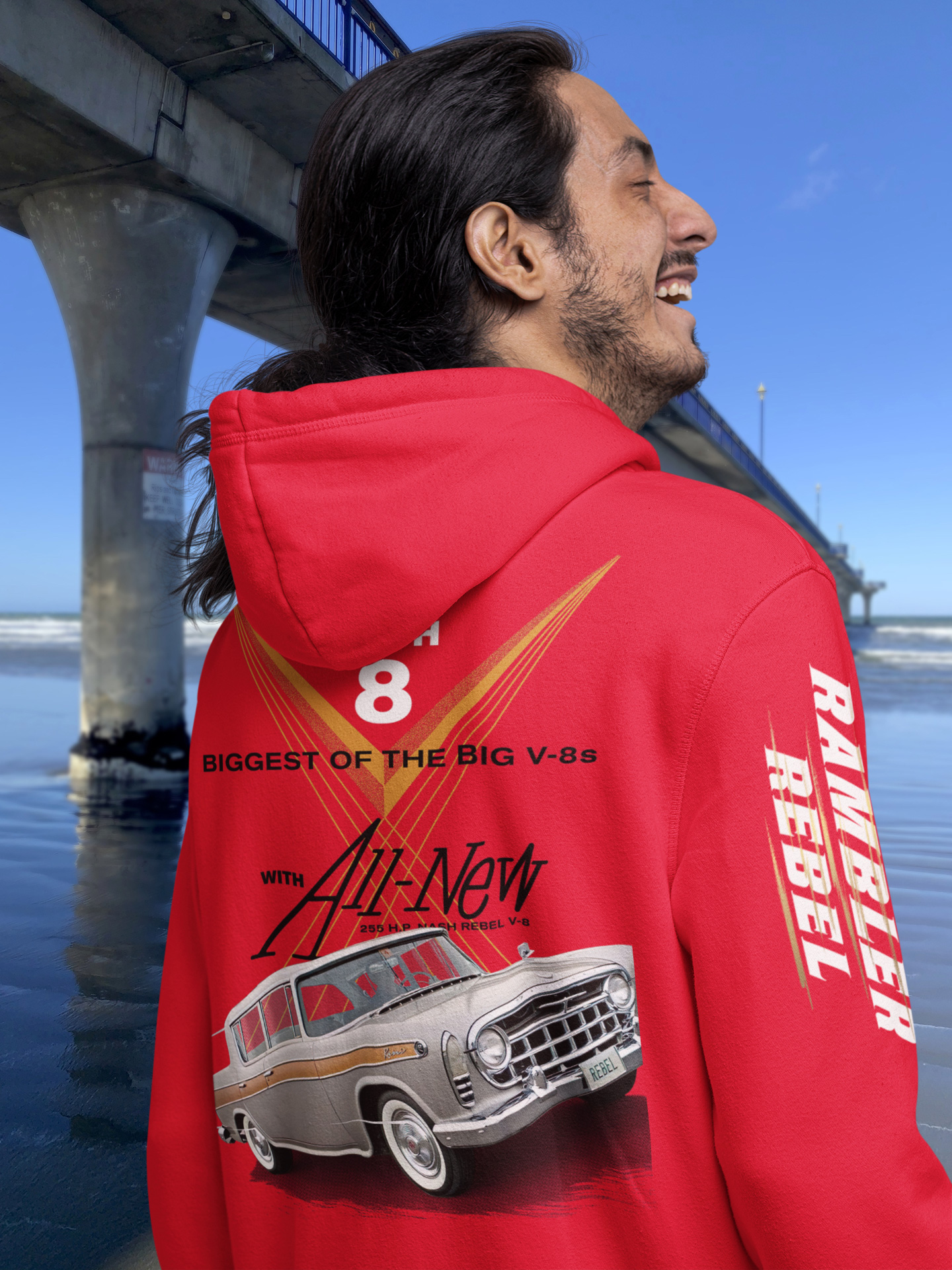Back view of a young man in a red hoodie with a 1957 Rambler Rebel print, featuring the car in motion on a test track, modelled beside Brighton Pier.