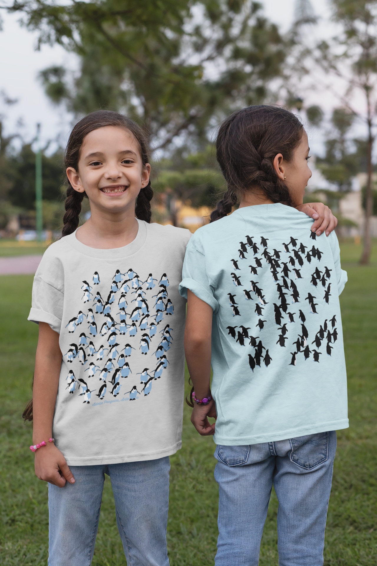 Twin girls in a park modeling a snow marle and a pale blue Adelie Penguin kids’ t-shirt, showing front and back views with a three-colour penguin.