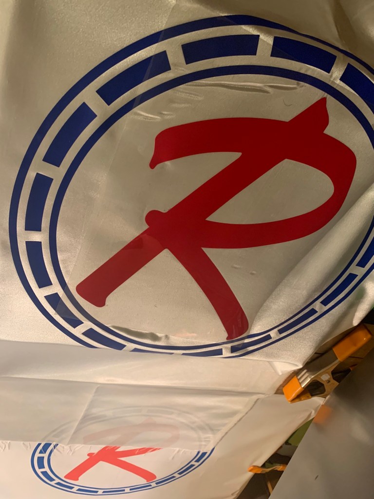 Image of a 1964 red, white, and blue Rambler banner on a heat press platen during a two-colour heat-pressing process in a workshop.
